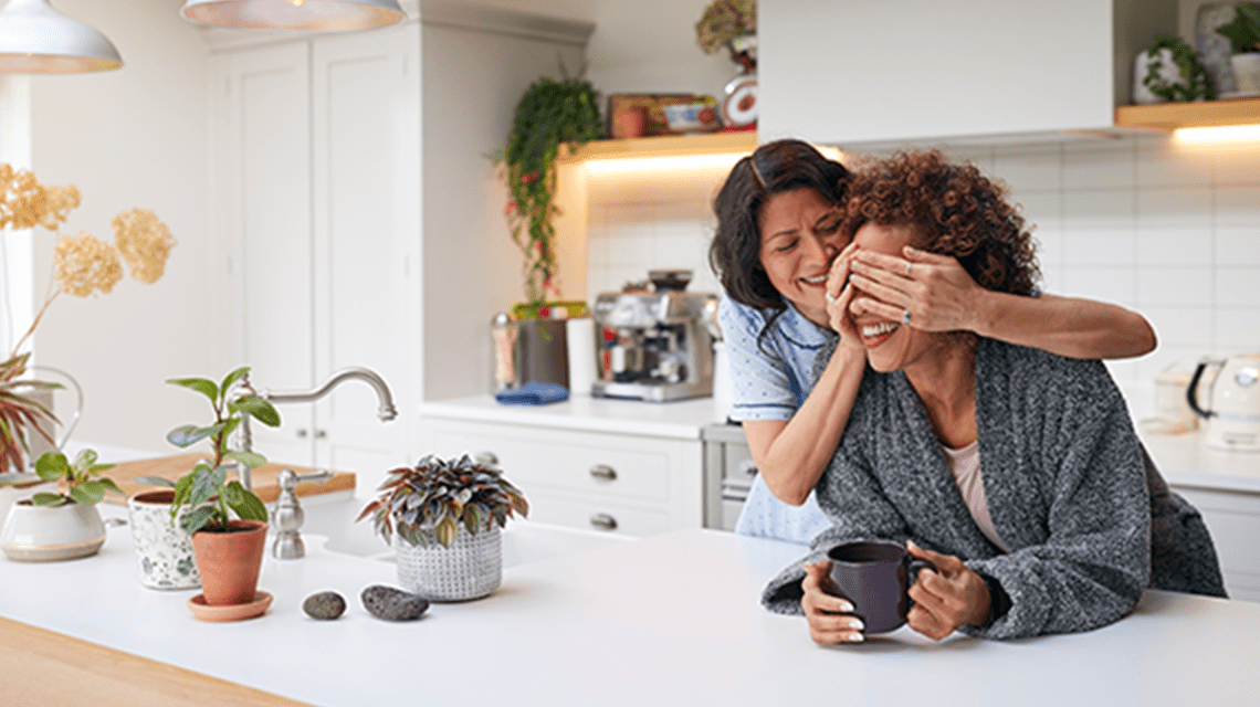 Couple in a modern white kitchen, one person is leaning on an open countertop while the other person is covering their eyes with their hands as if hiding a surprise